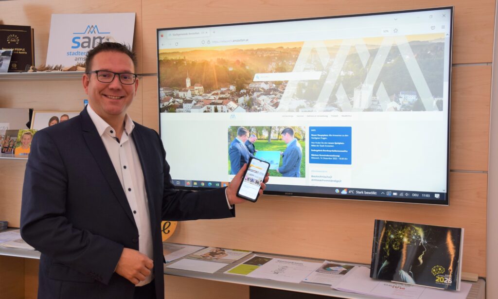 Bürgermeister Christian Haberhauer hält ein Handy mit der neuen Website in der Hand. Auf dem Bildschirm im Hintergrund ist ebenfalls die neue Webseite zu sehen.