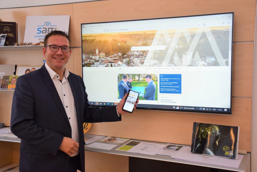 Bürgermeister Christian Haberhauer hält ein Handy mit der neuen Website in der Hand. Auf dem Bildschirm im Hintergrund ist ebenfalls die neue Webseite zu sehen.