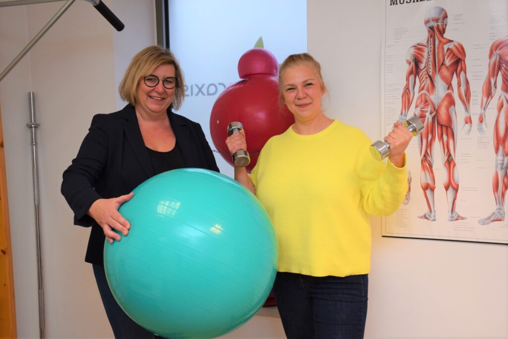 Bild von Claudia Weinbrenner und Birgit Huber. Frau Weinbrenner hält einen Gymnastikball und Frau Huber hält in jeder Hand eine Hantel.