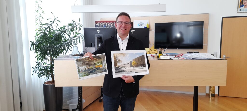 Bürgermeister Christian Haberhauer hält vor einem Stehschreibtisch zwei Bilder des neuen Hallenbades in den Händen