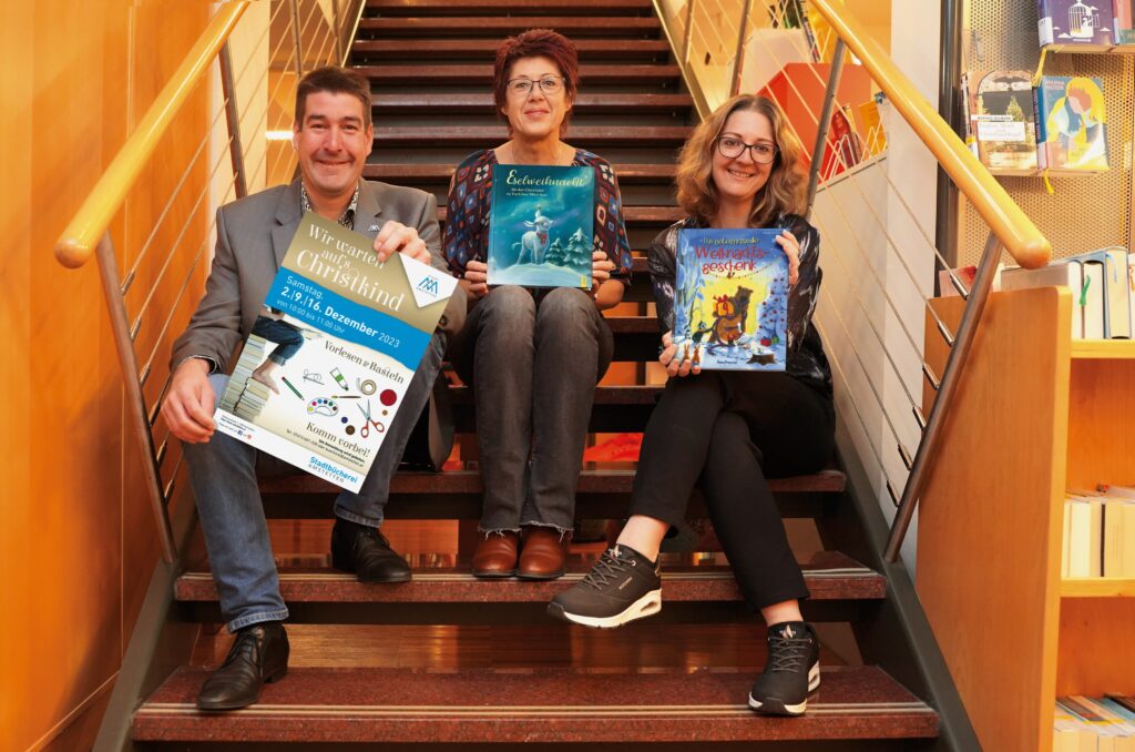 Kulturstadtrat Stefan Jandl, Rafetzeder Karin (Bücherei) und Schuster Marlene (Bücherei) sitzen auf der Büchereistiege und präsentieren Weihnachtsbücher und Plakat