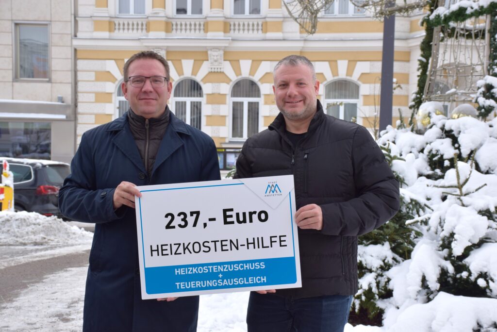 Bürgermeister Christian Haberhauer mit Vizebürgermeister Dominic Hörlezeder stehen mit einem Schild beschriftet mit 237 Euro Heizkostenhilfe vor verschneiten Bäumen.